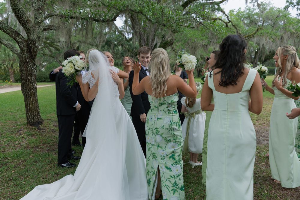 Wedding party gathers outdoors under oak trees, bride in white gown with veil, bridesmaids in green and white dresses.