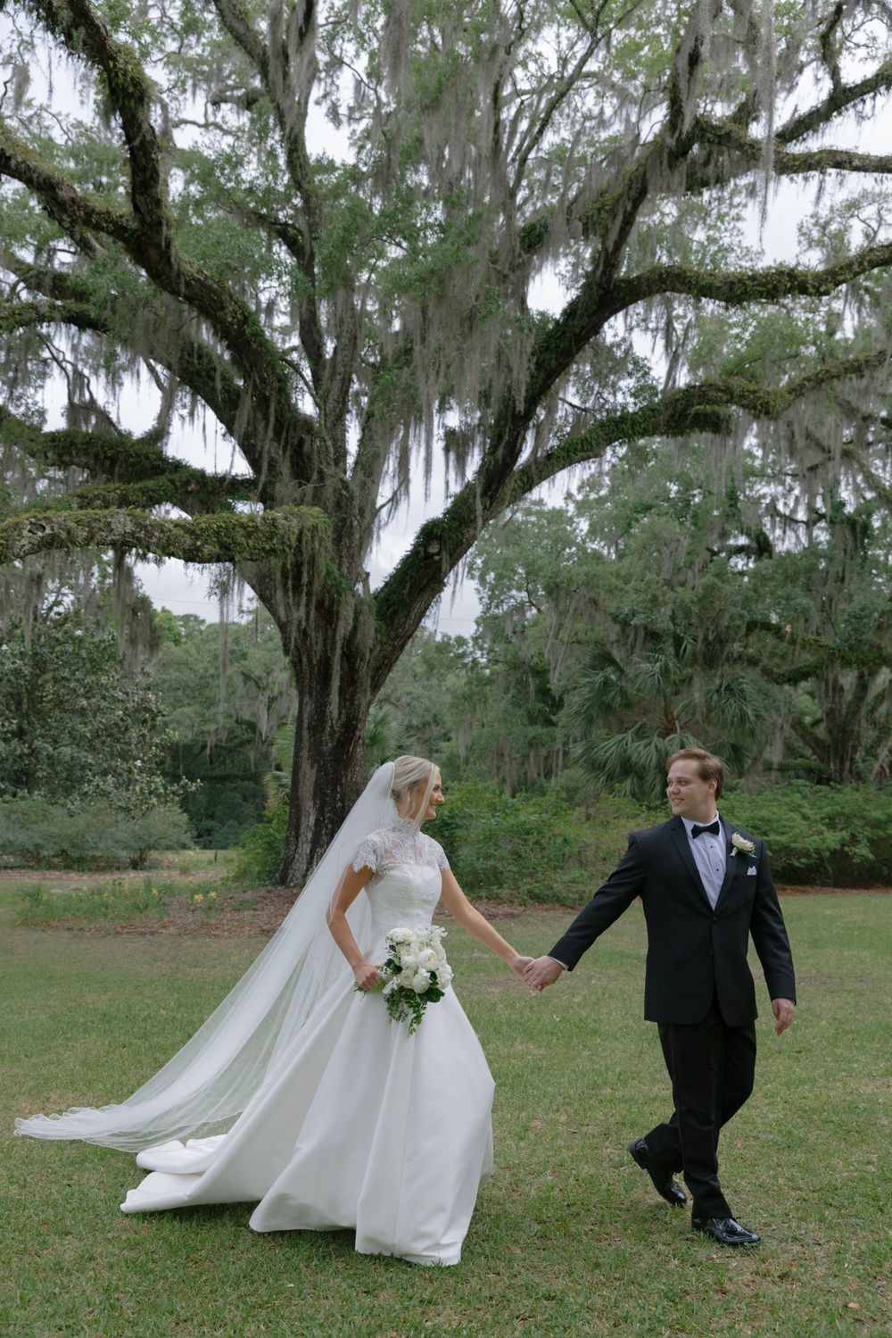 Bride and groom hold hands walking under a grand Spanish moss-draped oak tree on lush green grounds.
