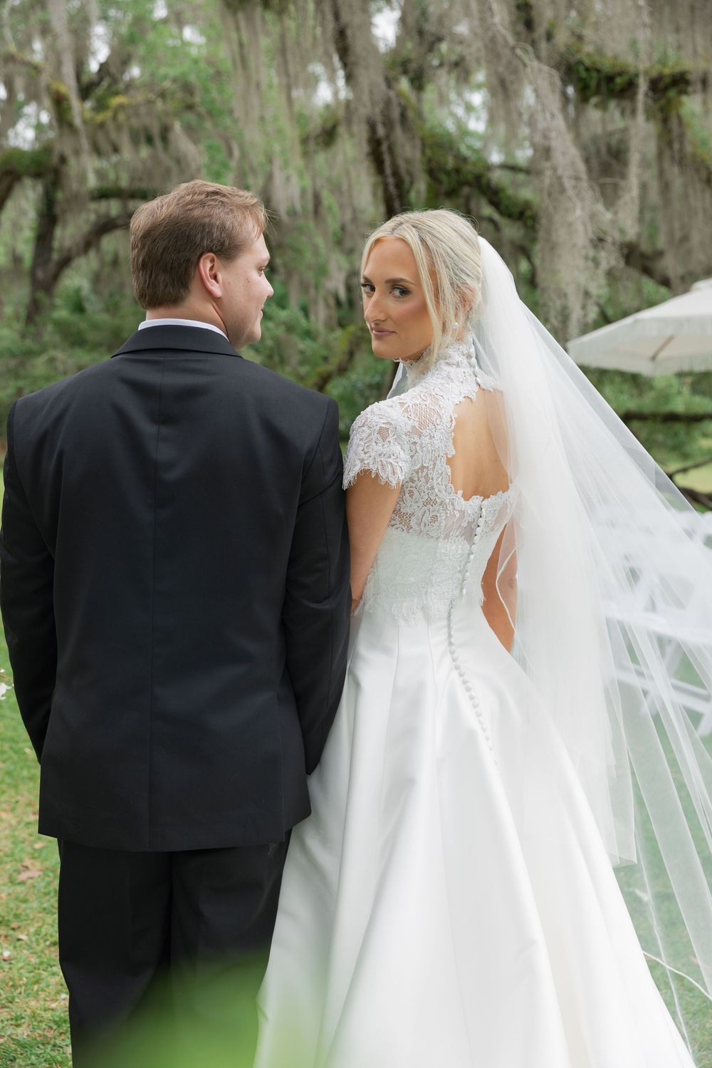 Bride in lace backless wedding gown with long veil stands beside groom outdoors under Spanish moss trees.