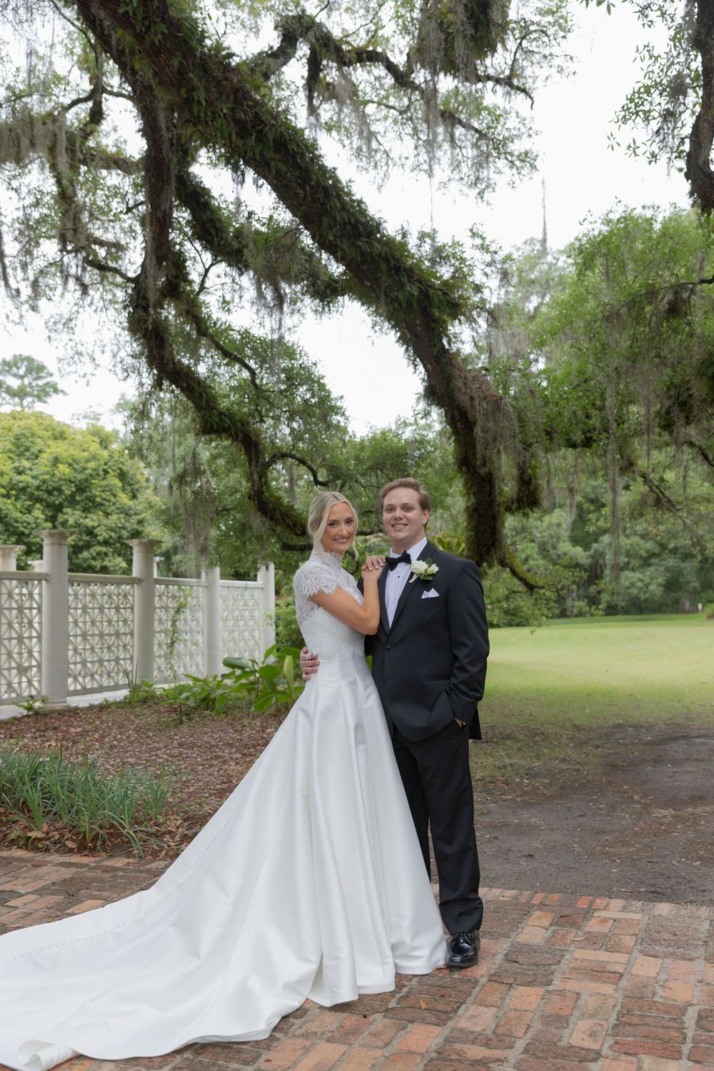 Bride and groom pose under Spanish moss-draped oak trees on brick path at elegant outdoor wedding venue.