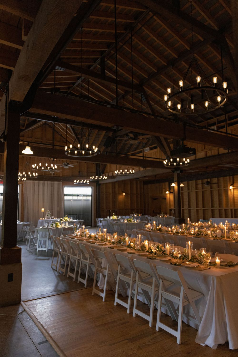Rustic barn wedding reception with long tables, white chairs, candlelight, and warm Edison bulb chandeliers hanging from wooden beams.