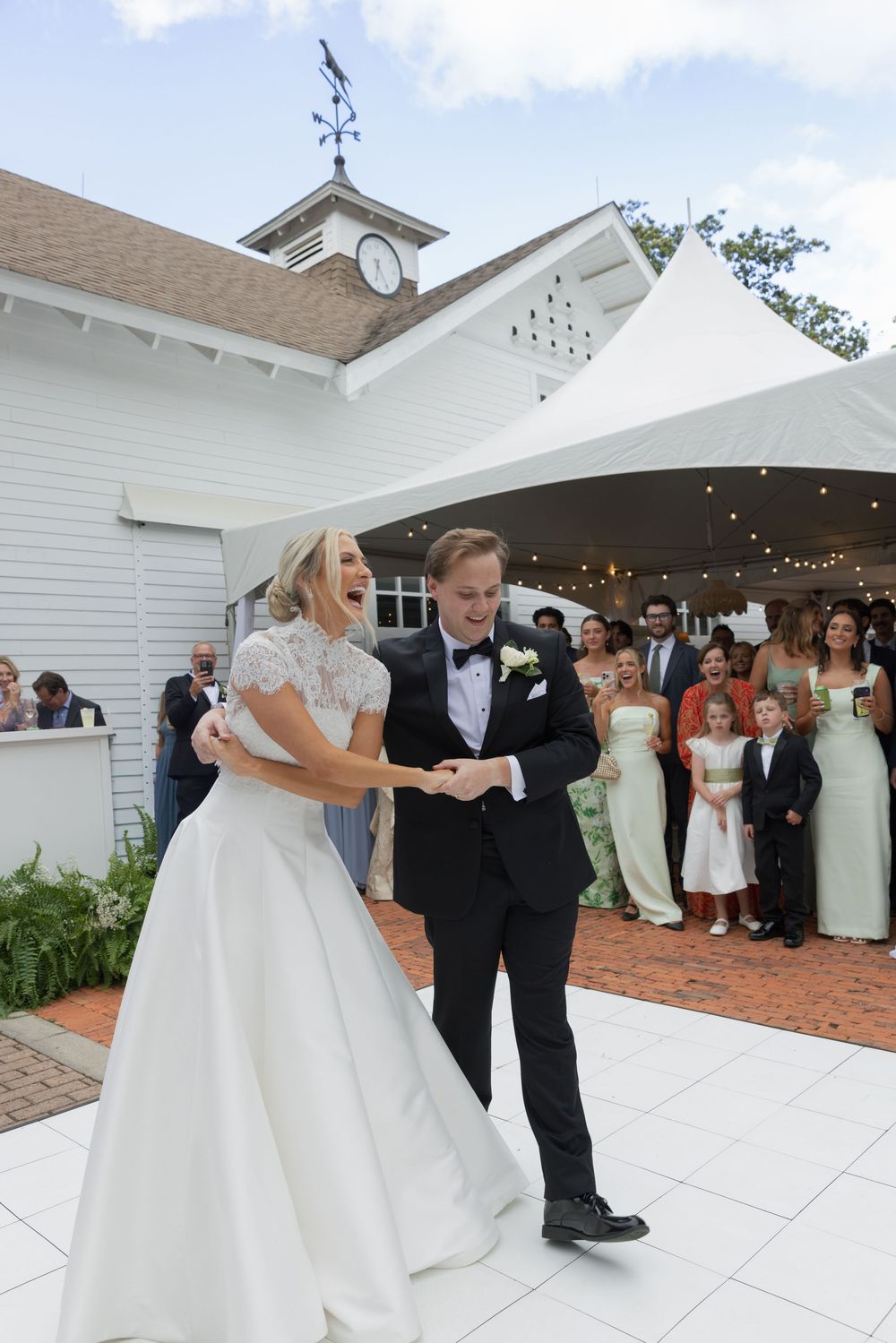Bride and groom share a joyful first dance outdoors at elegant white wedding venue with guests watching.