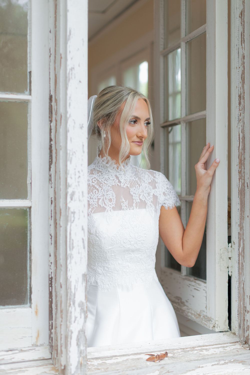 Elegant bride in lace high-neck wedding gown standing in vintage white doorway looking to the side.