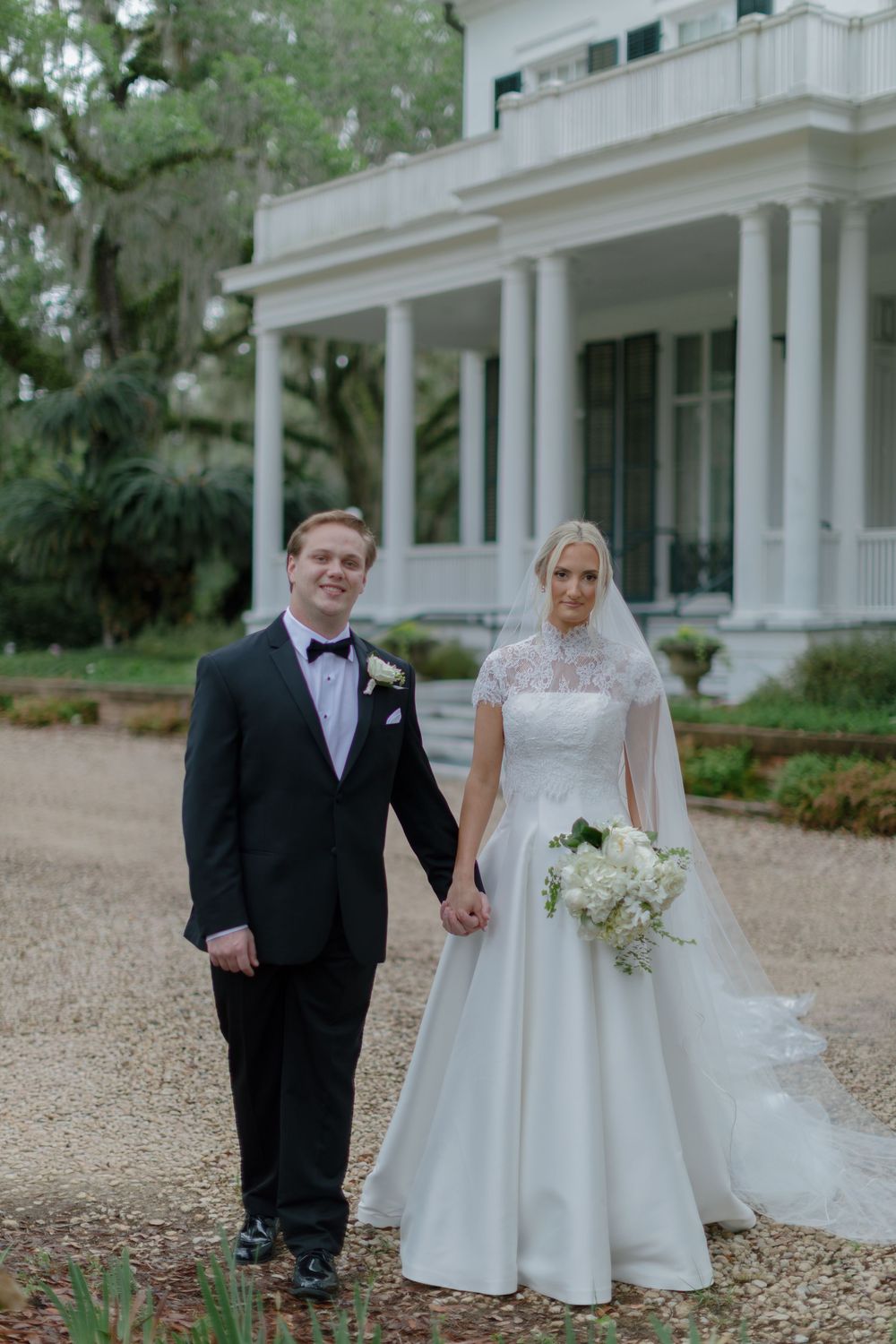 Bride and groom pose holding hands in front of elegant white Southern plantation home with columned porch.