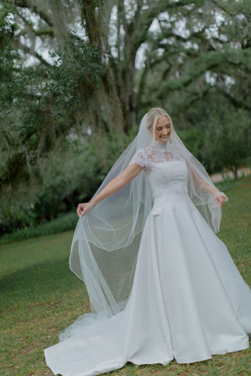 Bride in lace cap-sleeve gown twirls her cathedral veil beneath a large oak tree on green lawn.