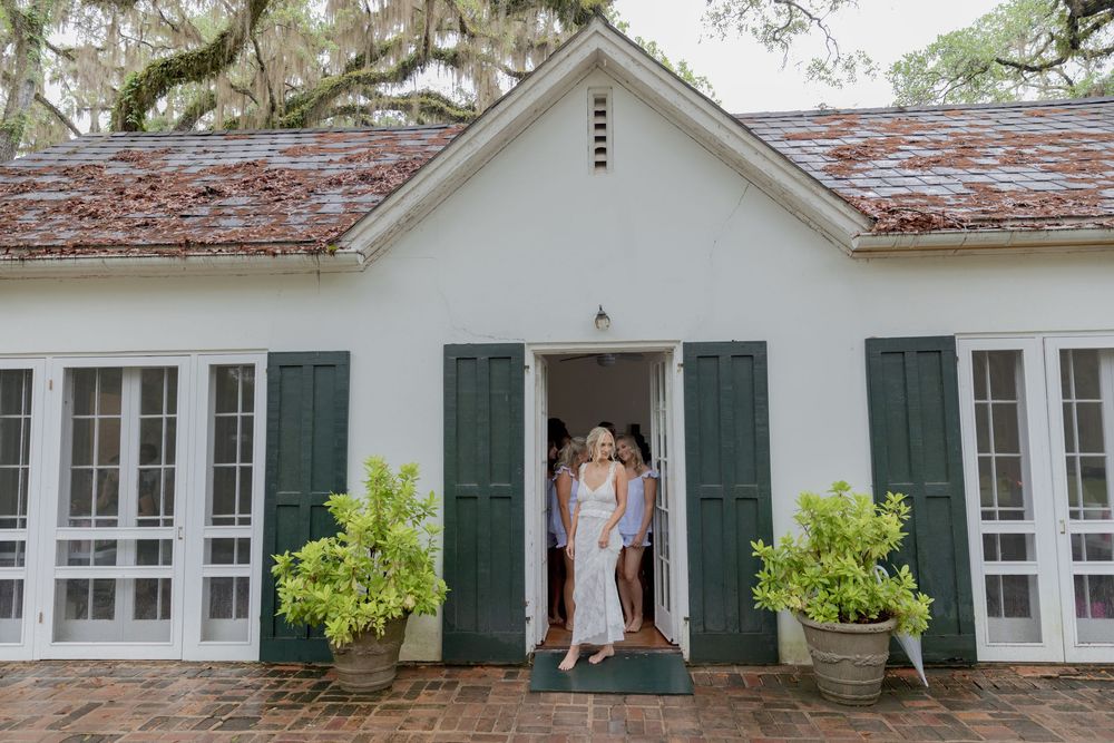 Two women stand in the doorway of a white cottage with green shutters, surrounded by lush potted ferns.