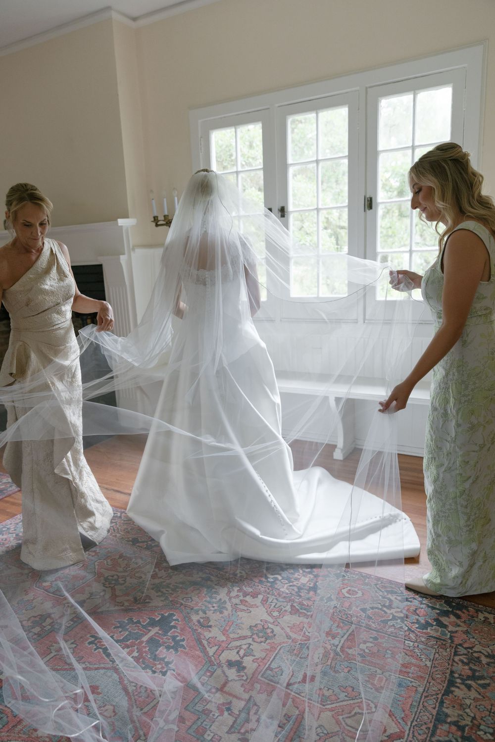 Two women help bride arrange long cathedral veil and train in bright interior room before wedding ceremony.