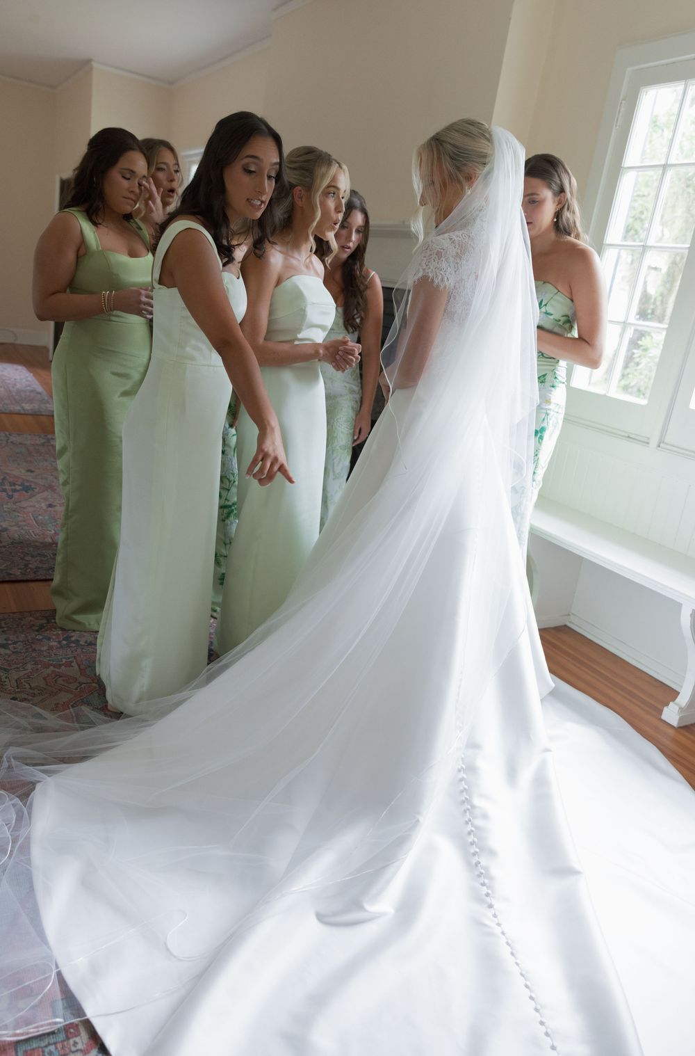 Bridesmaids in sage green gowns helping bride with long cathedral veil train indoors before ceremony.