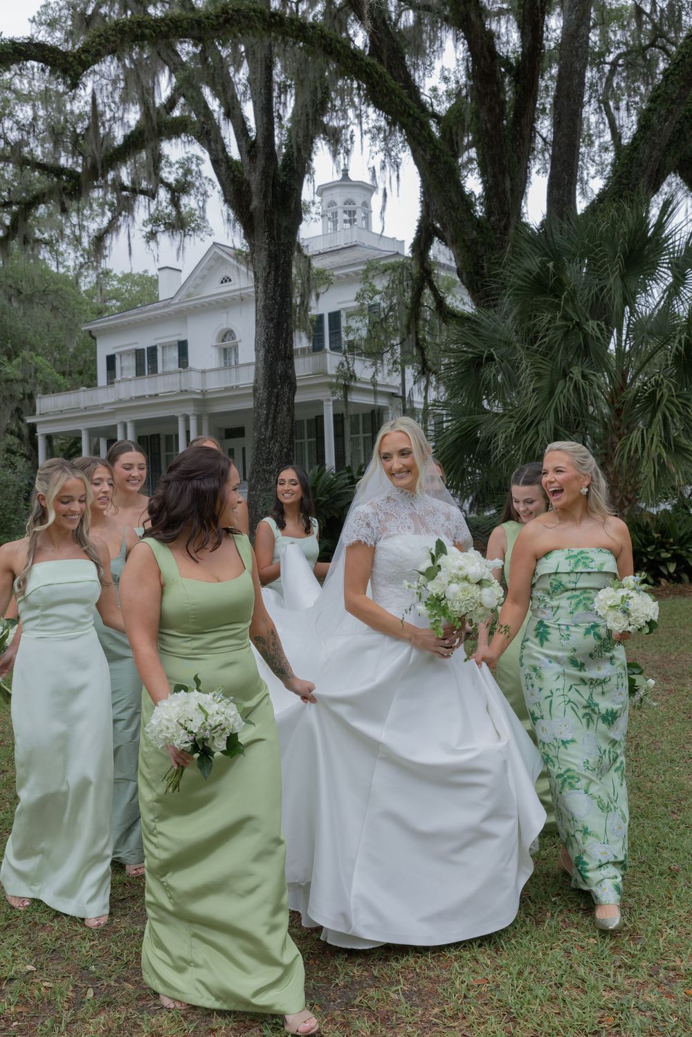 Bride in lace gown walks with bridesmaids in sage green dresses outside historic white Southern mansion.
