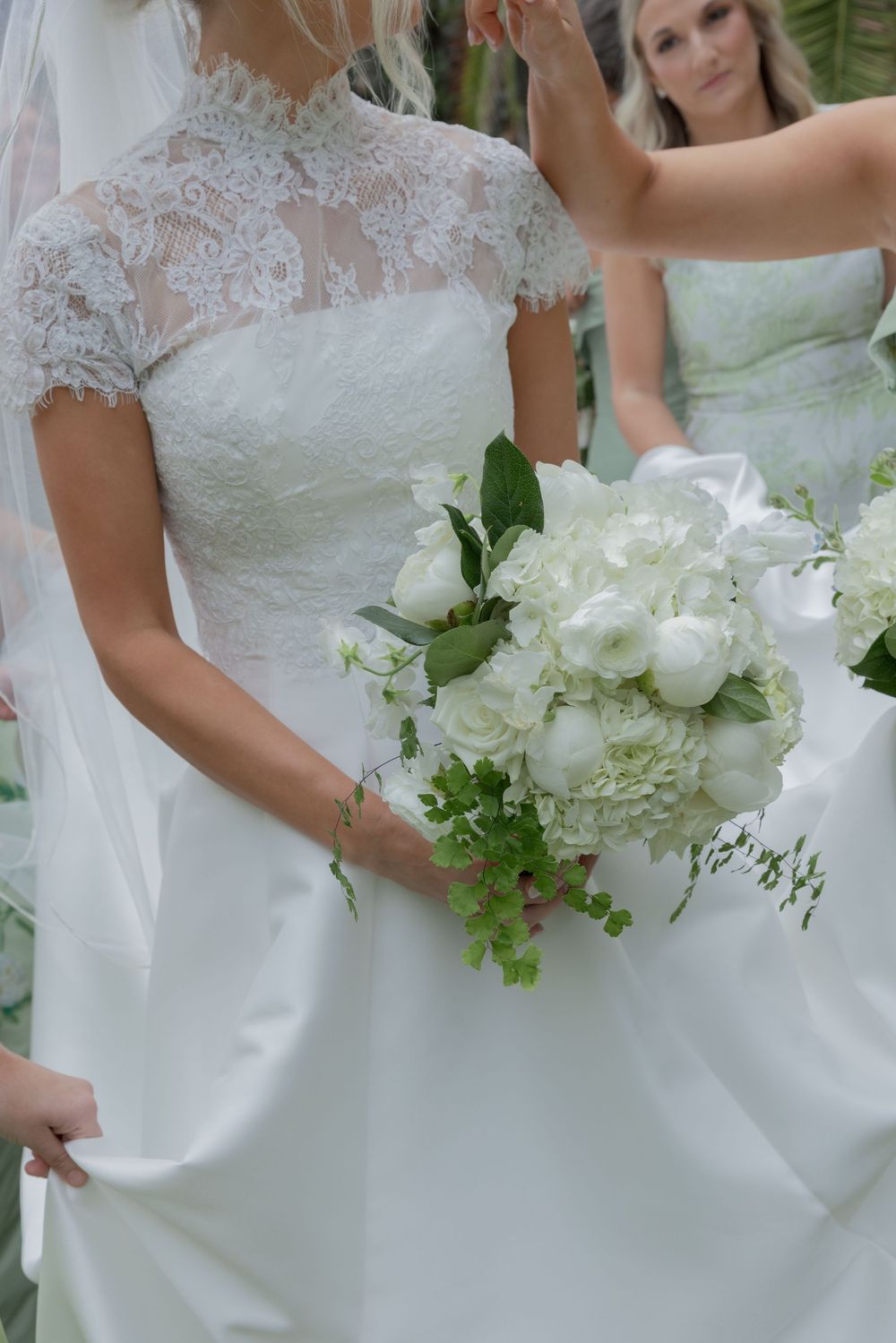 Bride in lace wedding gown holding white floral bouquet with greenery, bridesmaid adjusting veil in background.