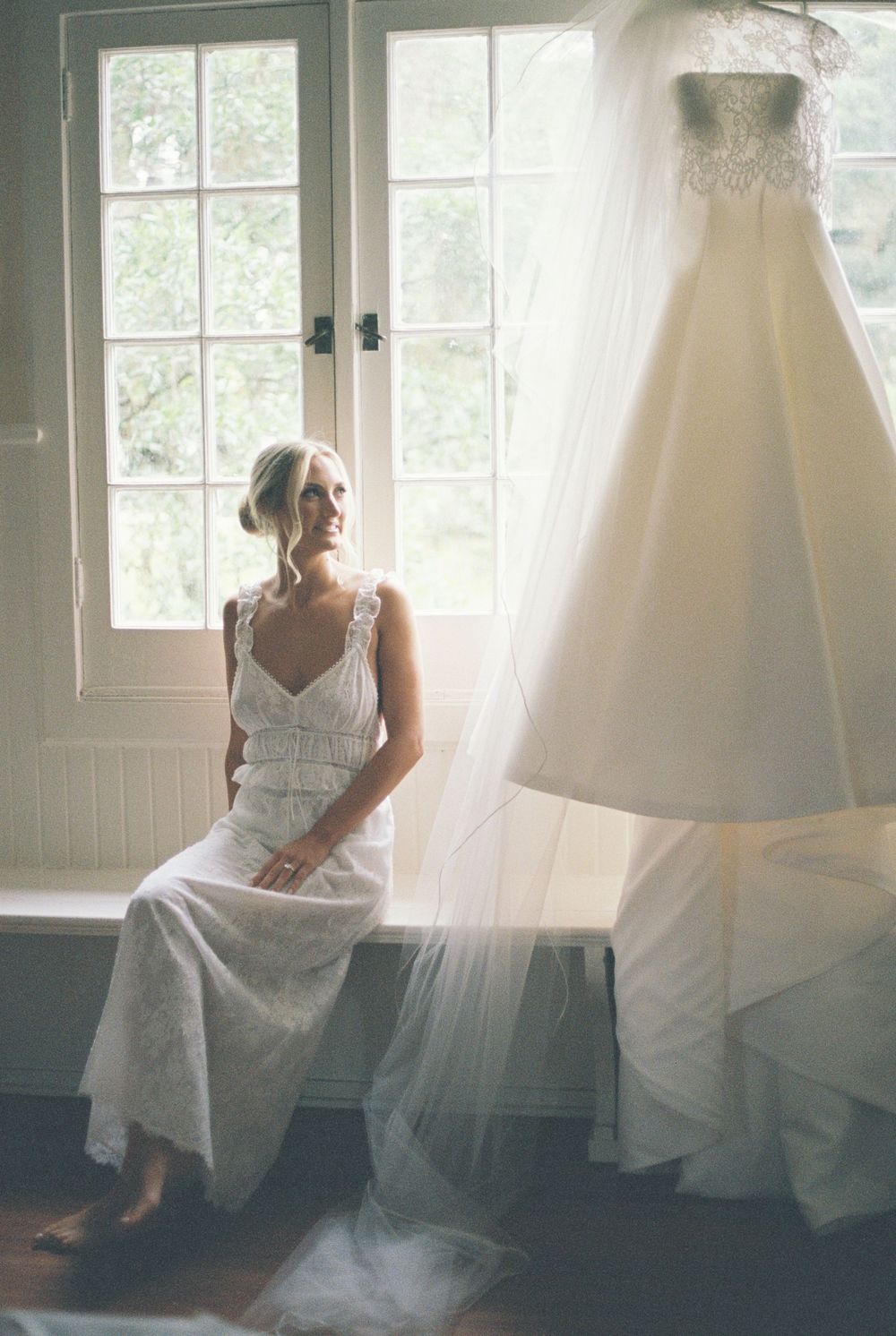 Blonde bride sits by window in white lace getting-ready outfit beside her hanging wedding gown with veil.