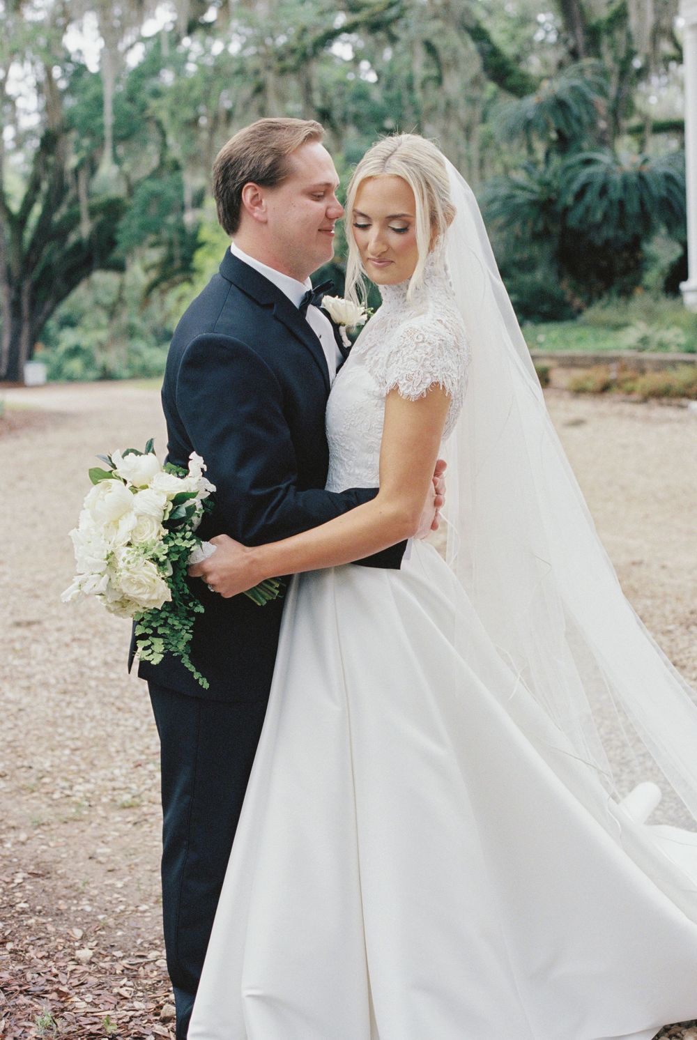 Bride and groom embrace tenderly outdoors under moss-draped trees, bride holding white floral bouquet.