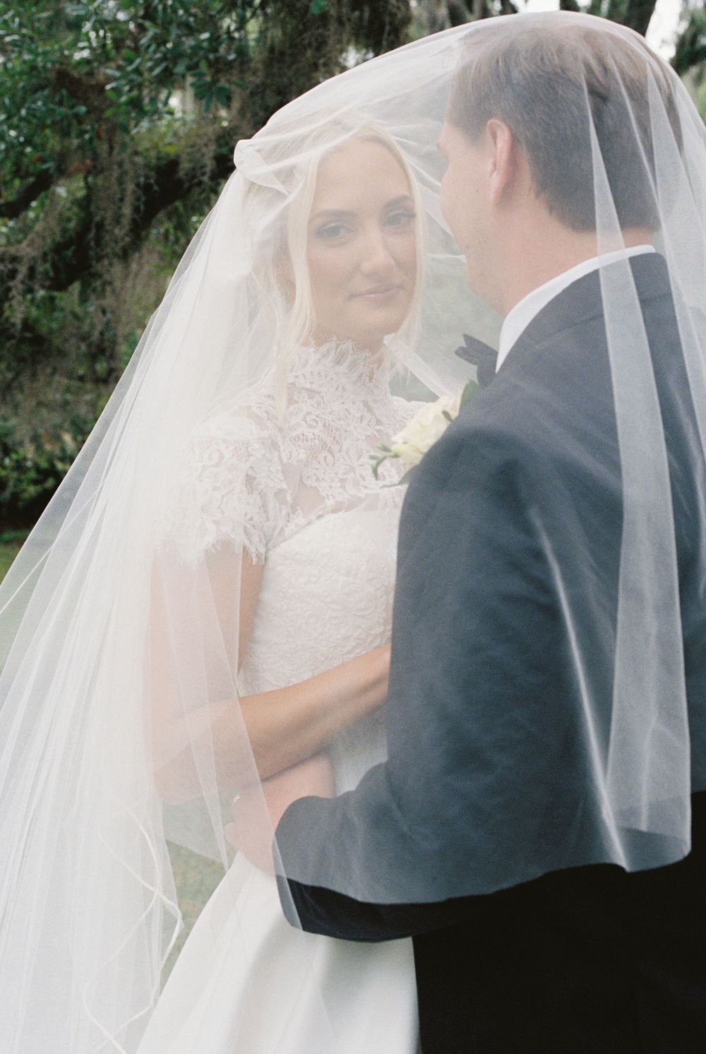 Bride and groom share a romantic moment under a flowing white wedding veil outdoors.