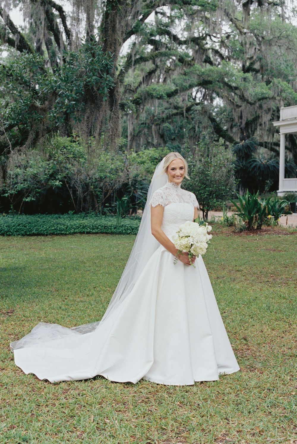 Bride in elegant lace ball gown with cathedral veil holding white bouquet outdoors under Spanish moss-draped oak trees.