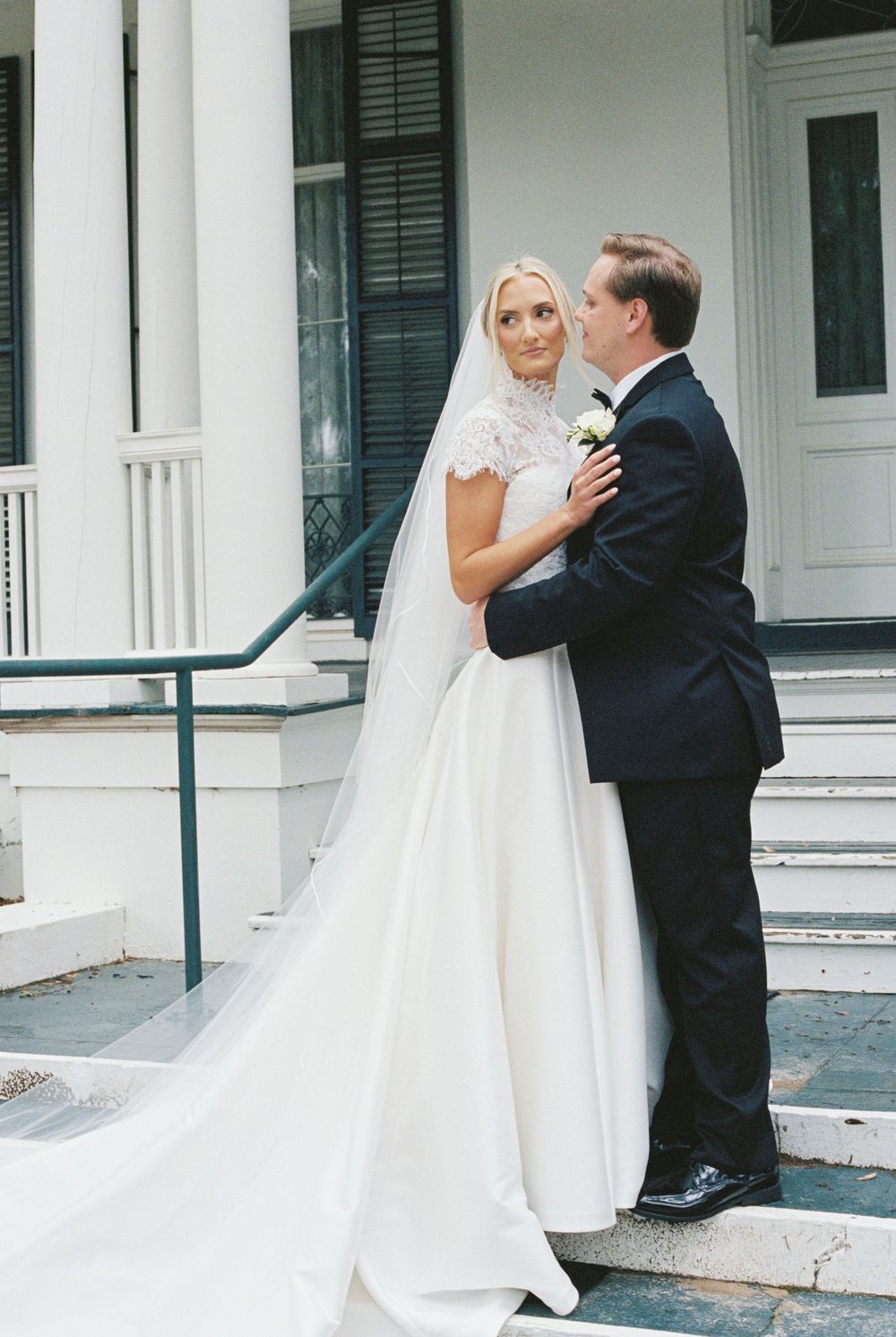 Bride and groom share romantic moment on steps of white Southern colonial mansion, bride in lace gown.