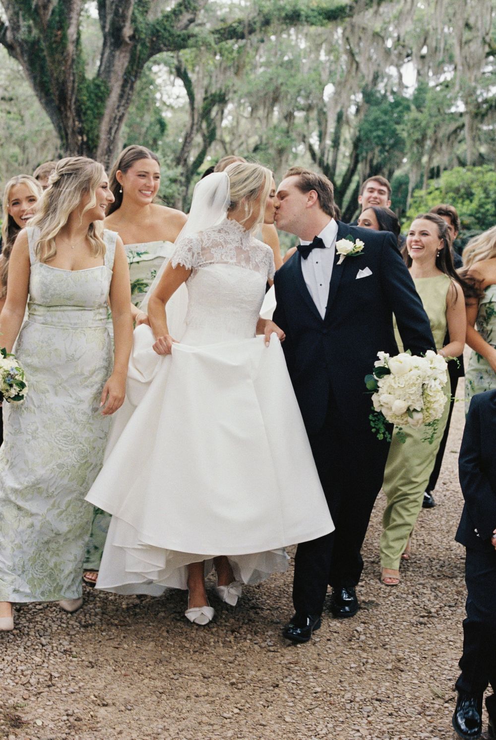 Bride and groom kiss while walking with smiling wedding party in green dresses along tree-lined path.
