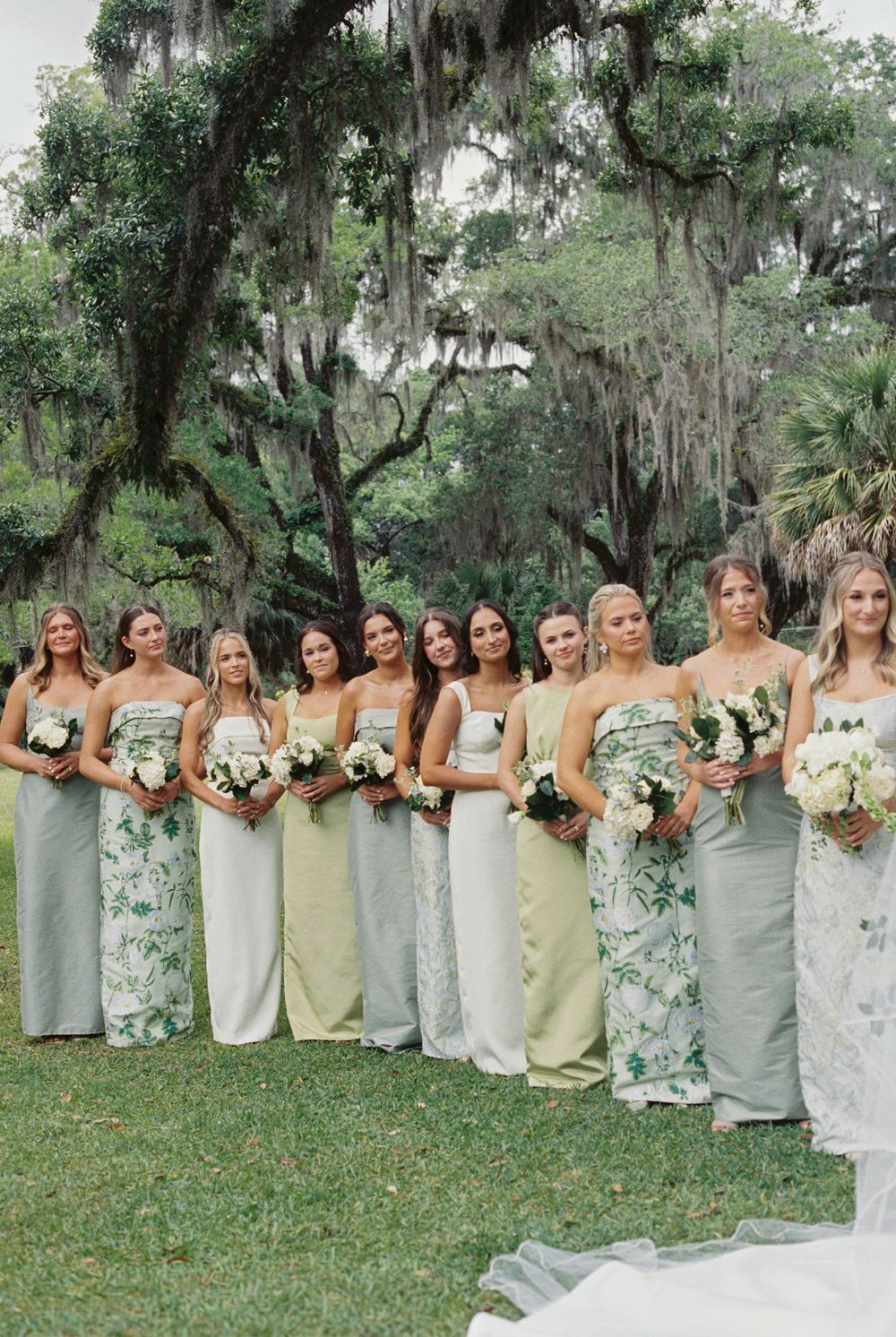 Large bridal party in mixed sage green and floral gowns holding white bouquets under Spanish moss trees.