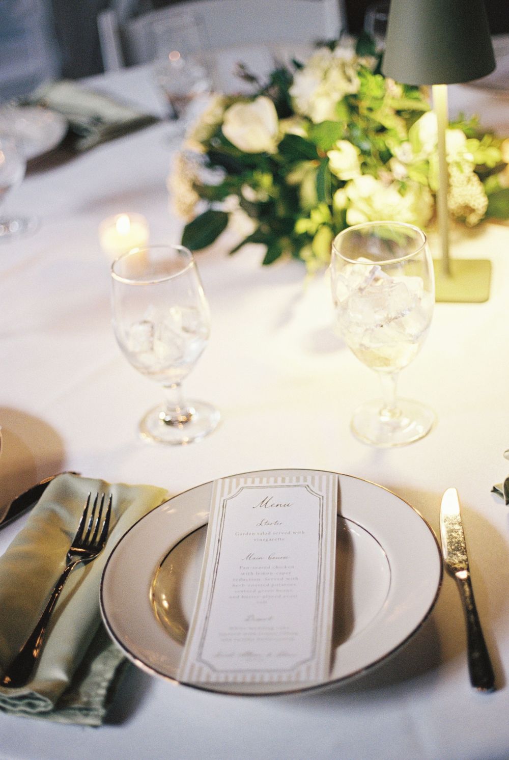 Elegant wedding reception place setting with white plates, menu card, wine glasses, and floral centerpiece.