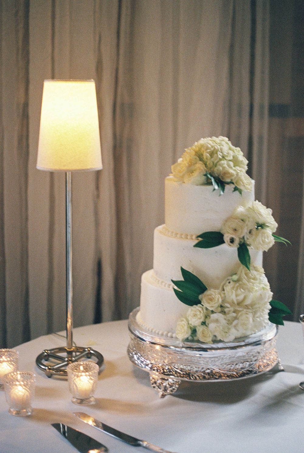 Elegant three-tier white wedding cake adorned with white roses and hydrangeas on silver stand beside glowing lamp.