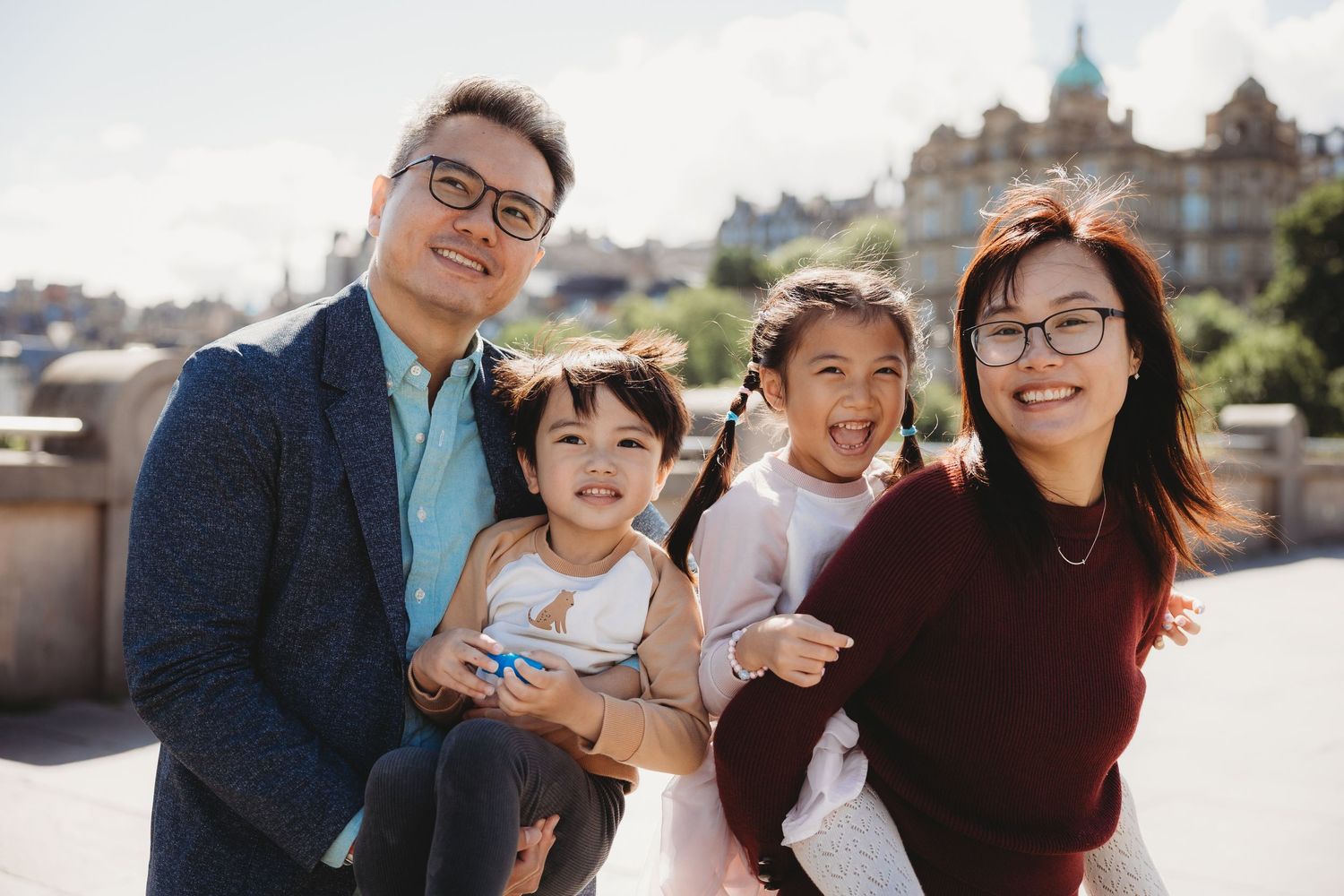Happy Asian family of four posing outdoors with historic buildings in background on a sunny day.