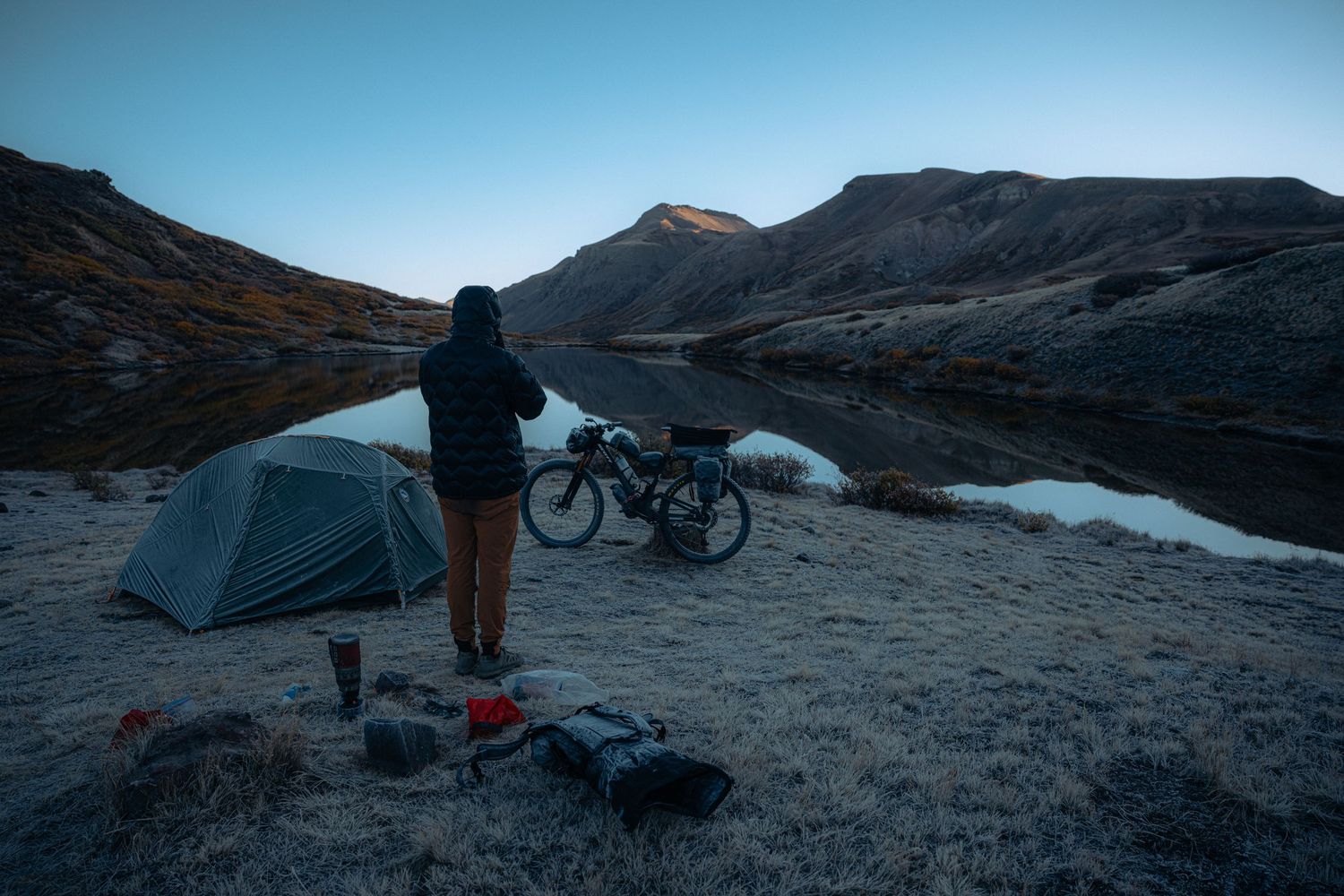 Person camping by a serene mountain lake at dusk with a tent, bicycle, and gear on a frosty alpine shoreline.