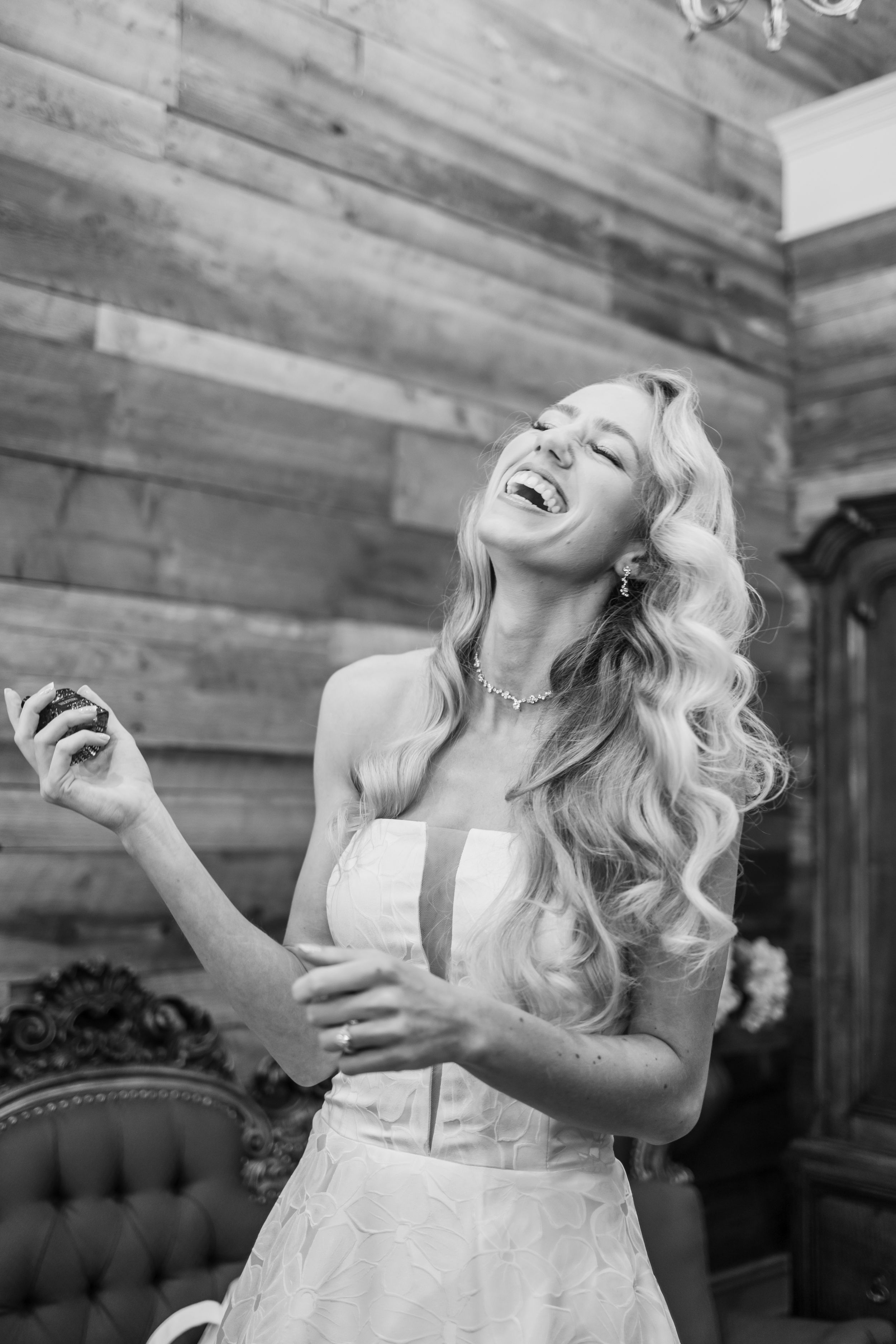 Black and white photo of a laughing blonde bride in a strapless gown holding perfume, with long wavy hair.