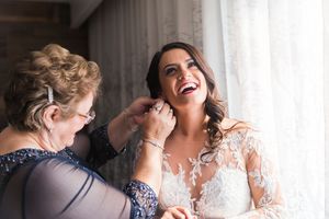 Bride laughing joyfully as older woman helps fasten her lace wedding dress near a bright window.