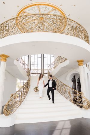 Bride and groom descend elegant white staircase with ornate gold railings in a grand wedding venue.