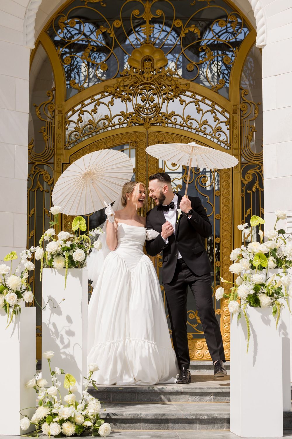 Bride and groom laughing under white parasols before ornate golden gates with white floral arrangements.