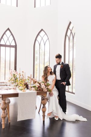 Bride and groom pose elegantly beside a floral wedding reception table in a bright chapel with gothic arched windows.