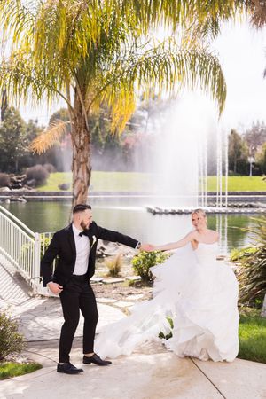 Bride in white gown and groom in black tuxedo dancing by a fountain and palm tree at an outdoor wedding venue.