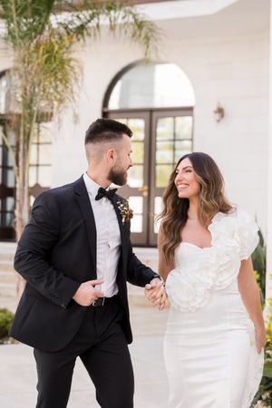 A smiling bride in a white floral gown holds hands with a groom in a black tuxedo outside an elegant venue.