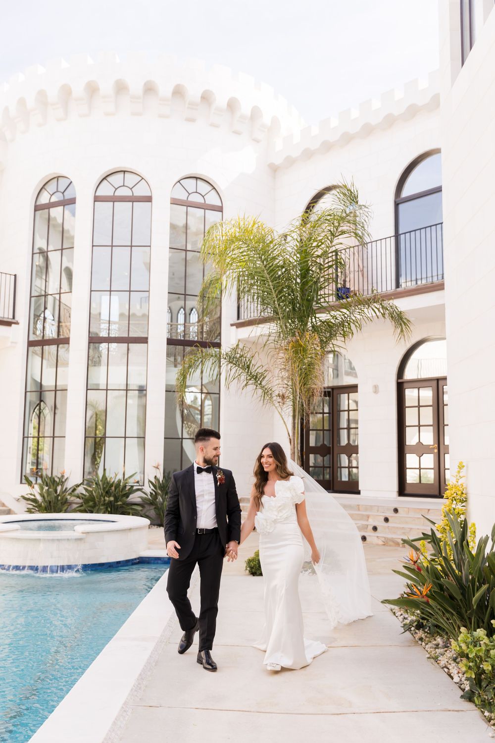Bride and groom walk hand-in-hand beside a pool at a white Spanish-style villa on their wedding day.