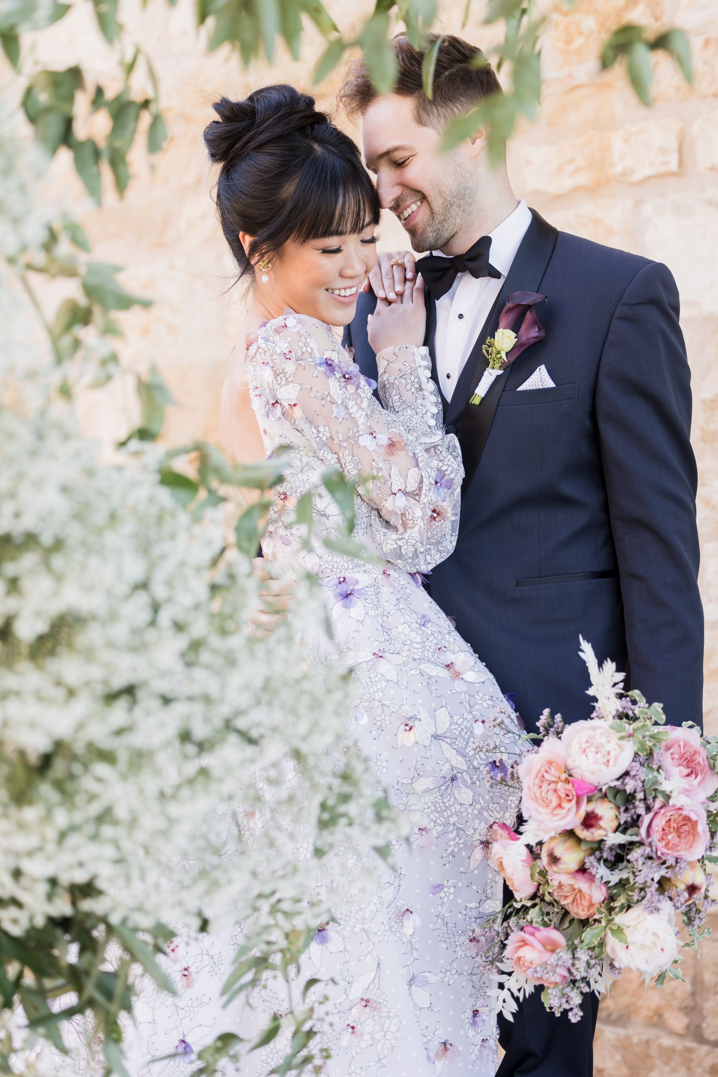A joyful bride in a beaded gown and groom in navy tuxedo share a romantic moment outdoors with a pink floral bouquet.