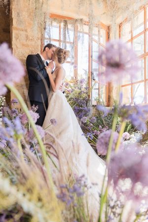 Bride and groom share a romantic kiss surrounded by purple wildflowers in a sunlit rustic venue.