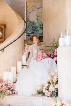 Bride in a beaded long-sleeve gown sits on floral-adorned stairs with pink roses and white candles.