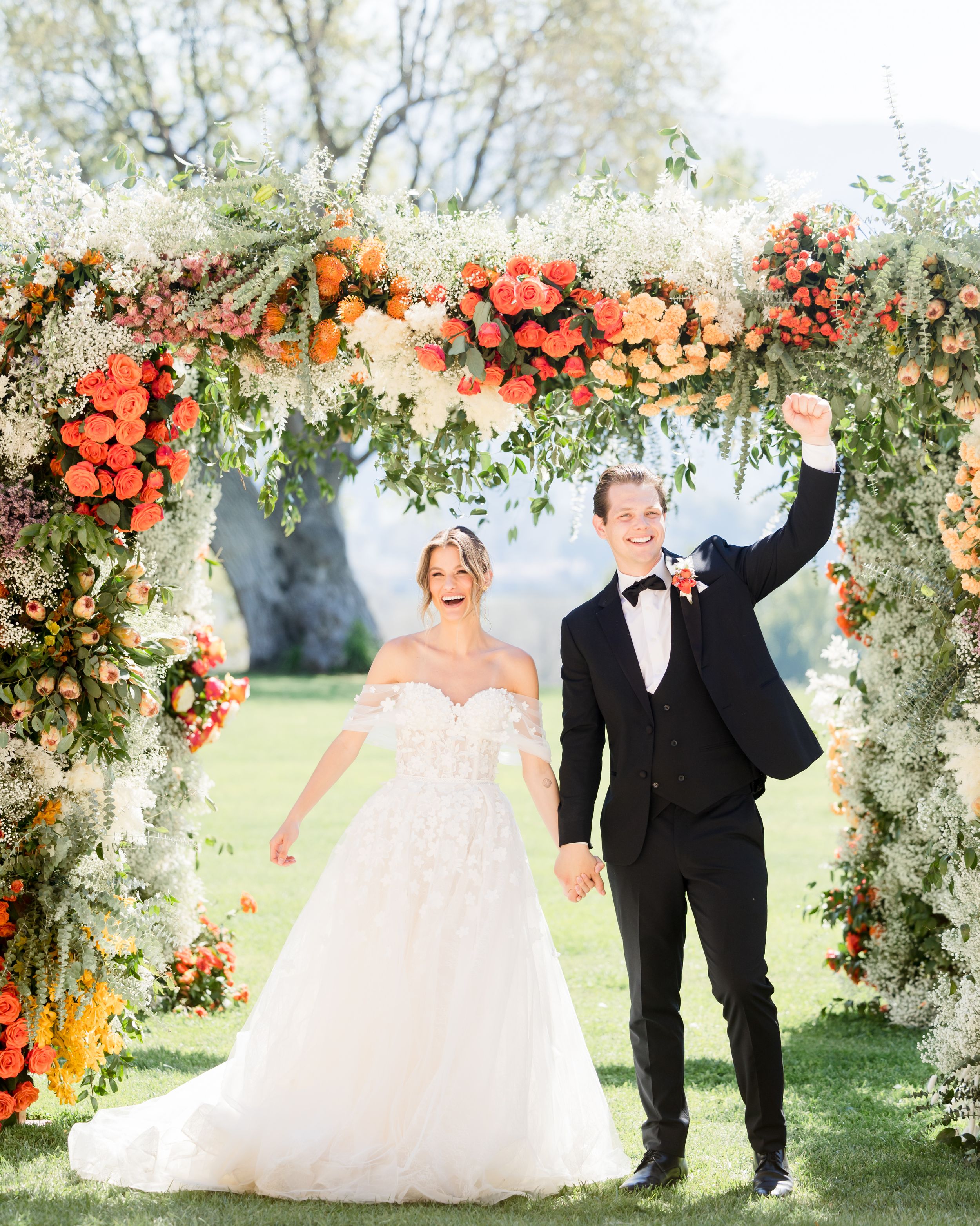 Bride and groom celebrating under a vibrant floral arch with orange and white blooms at an outdoor wedding ceremony.
