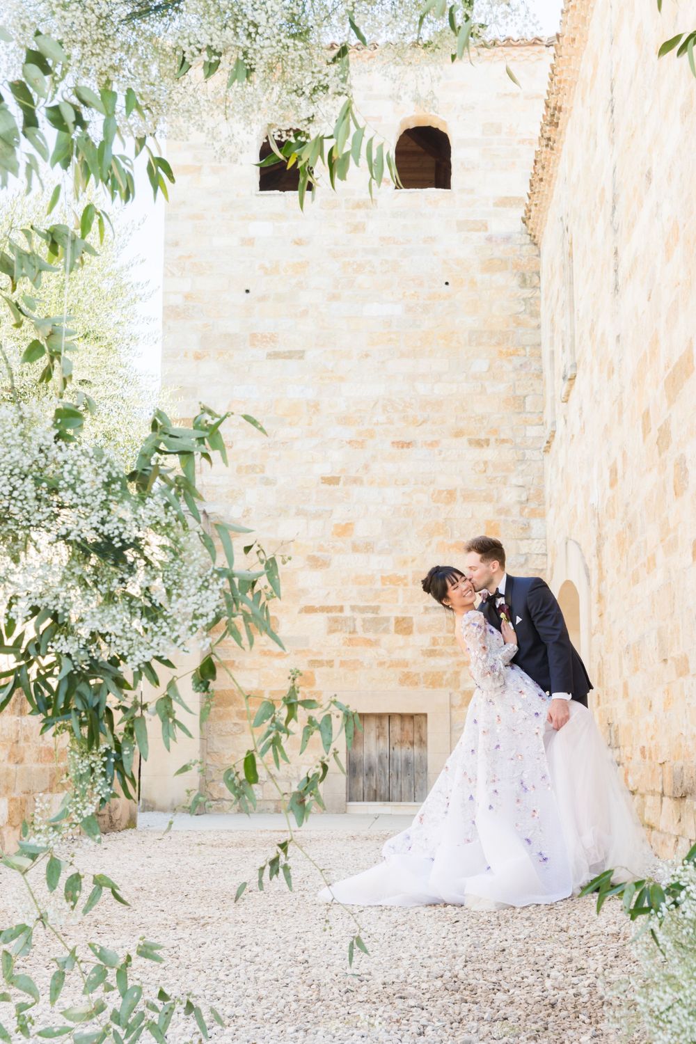 Bride and groom sharing a romantic kiss beside a rustic stone building surrounded by lush greenery and white florals.