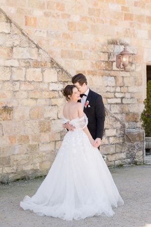Bride in white ball gown and groom in black tuxedo embrace romantically against a rustic stone wall.