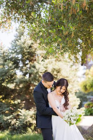 Bride and groom embracing outdoors under lush green trees, groom in black tuxedo, bride in white gown holding white floral bouquet.