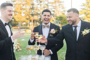 Three groomsmen in tuxedos laughing and toasting with champagne glasses at an outdoor wedding celebration.