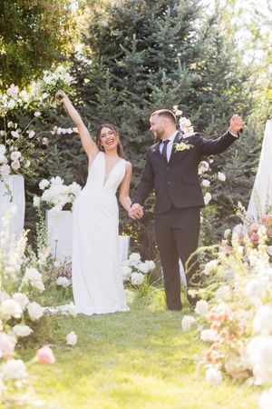 Joyful bride and groom celebrate after outdoor wedding ceremony, raising arms among floral arrangements.