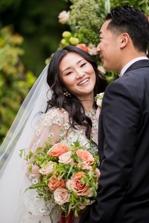 Smiling Asian bride in floral gown holding peach roses bouquet stands with groom in black suit at outdoor wedding ceremony.