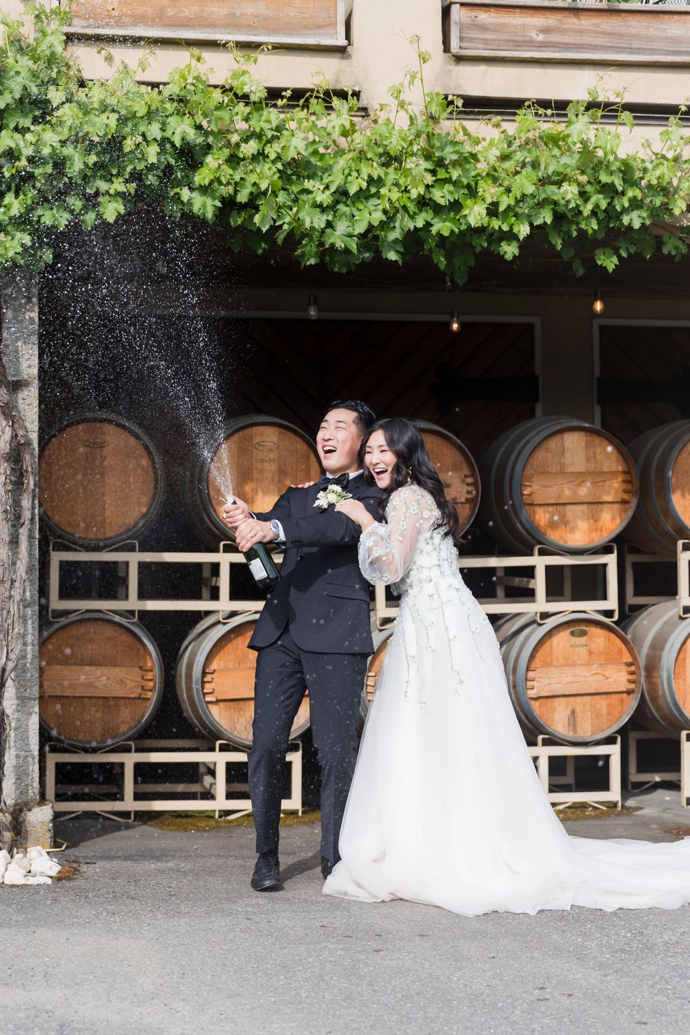 Joyful newlyweds popping champagne in front of wine barrels at a winery wedding celebration.