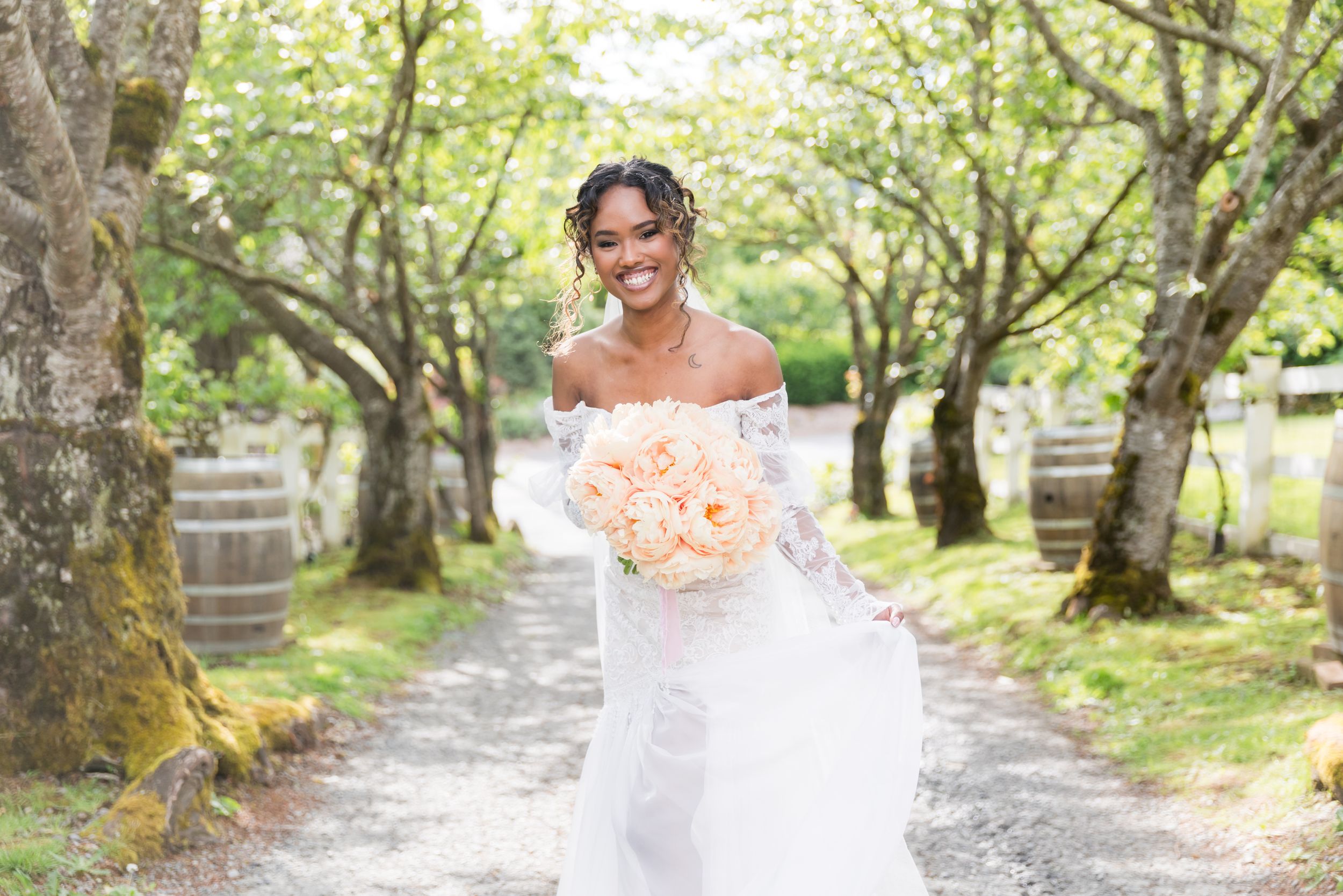 Smiling bride in white off-shoulder gown holding peach bouquet walking down tree-lined path with wooden barrels.