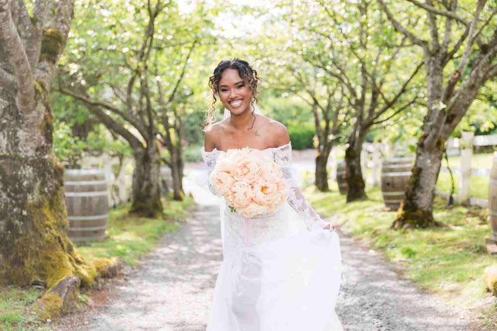 Smiling bride in white off-shoulder gown holding peach bouquet walking down tree-lined path with wooden barrels.