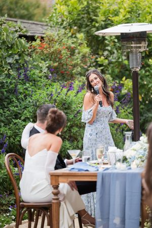 Woman in floral dress giving a speech at an outdoor wedding reception with lush garden backdrop.