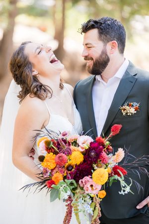 Laughing bride in white gown holds colorful bouquet beside smiling bearded groom in gray suit outdoors.