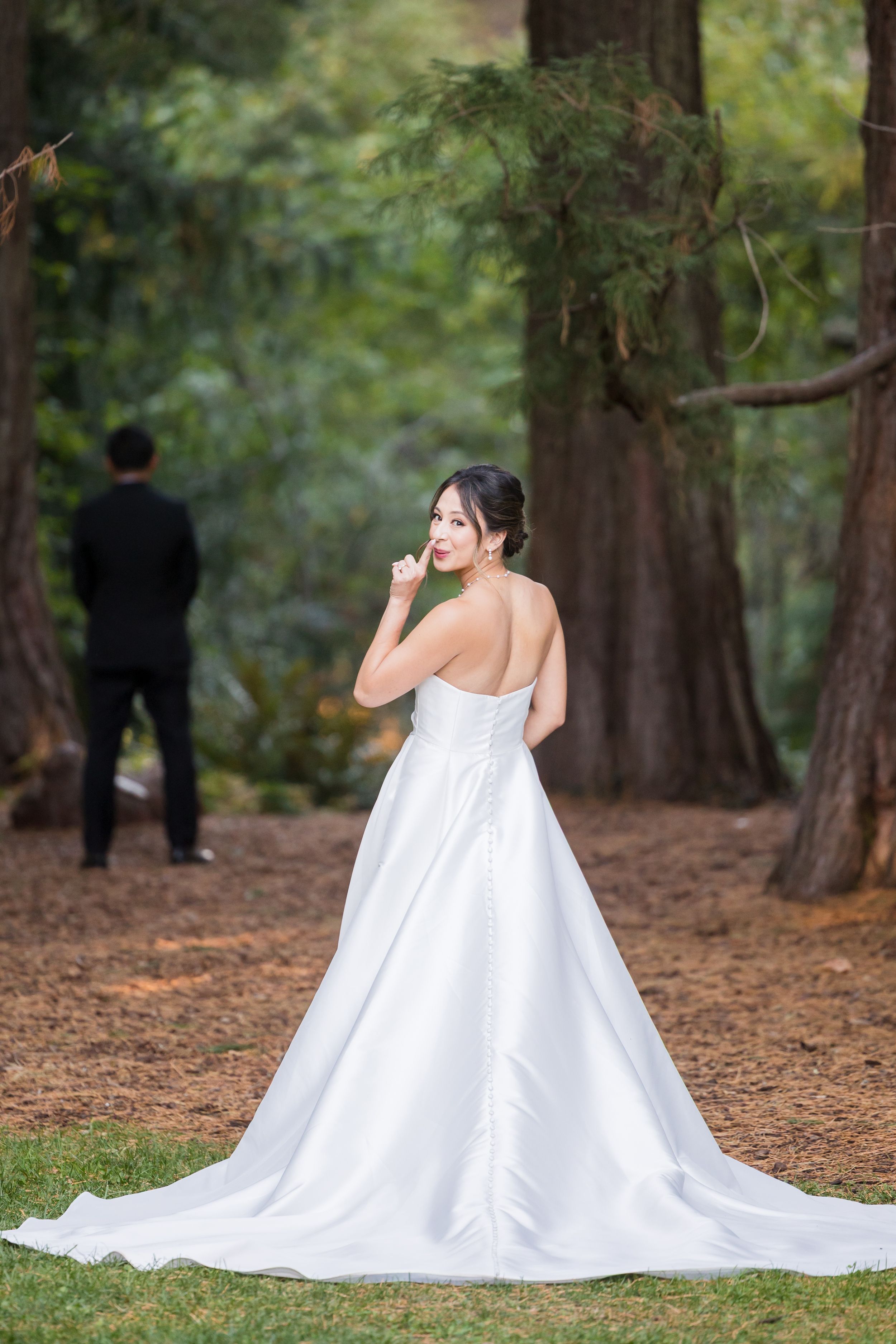 Bride in elegant white satin ballgown with train poses in forest, groom in black suit stands blurred in background.