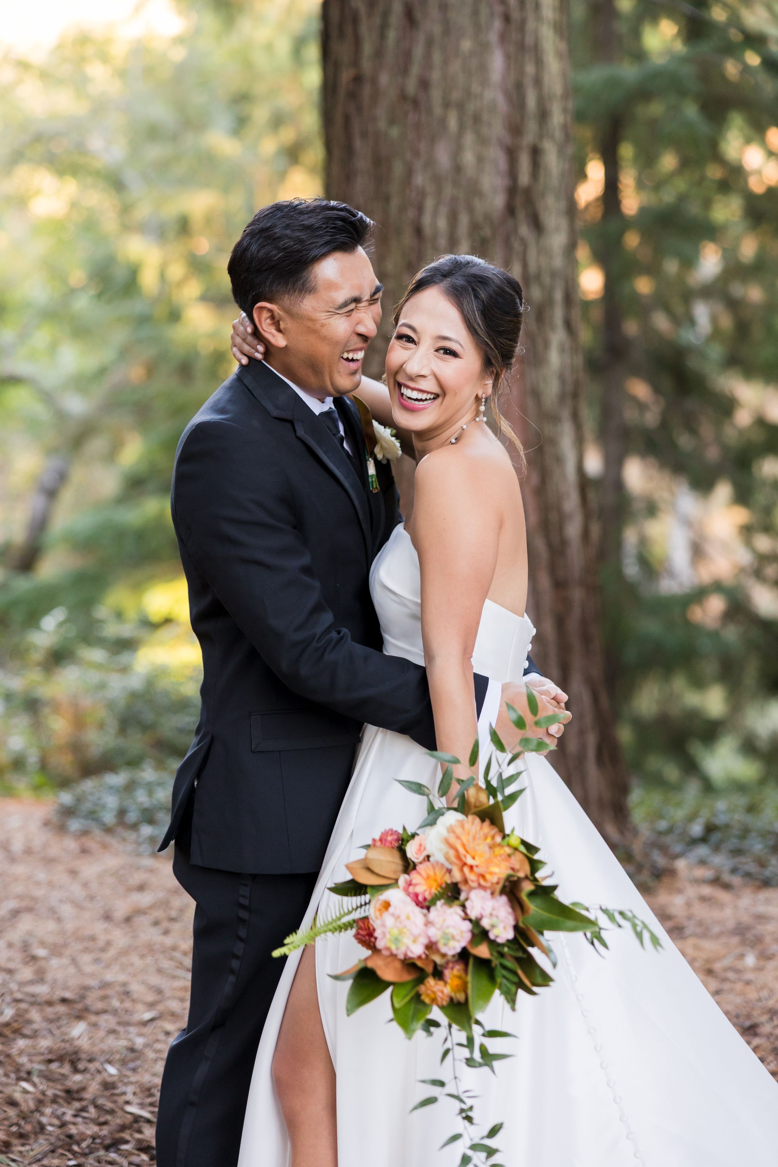 Bride and groom embracing in a redwood forest, bride holding a colorful bouquet of orange and pink flowers in a white gown.